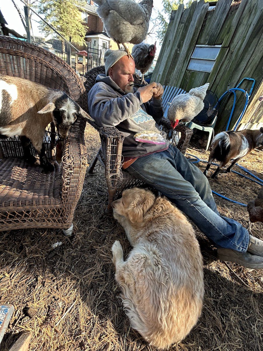 Al Curtis sitting outdoors with a rescue goat, chickens, and Toby the golden retriever at Legendary Acres.